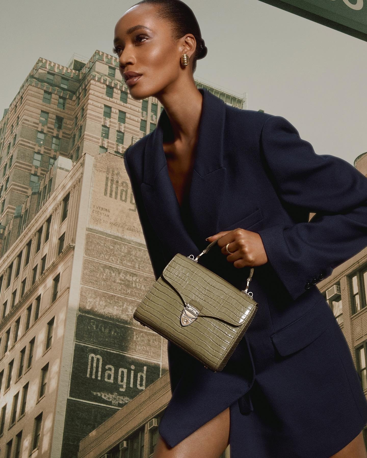 Model in dark blue blazer holding a textured olive green handbag against a tall city building background.