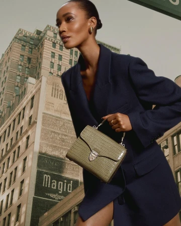 Model in dark blue blazer holding a textured olive green handbag against a tall city building background.