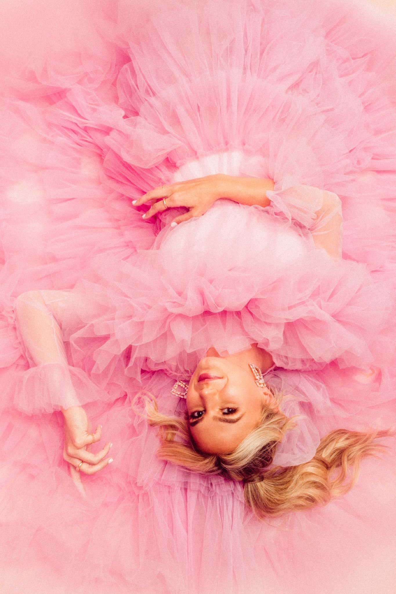 Woman in a voluminous pink tulle gown lies down. The gown is ruffled, with long sleeves, and the model wears statement earrings.