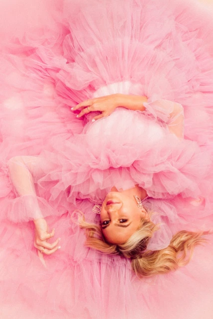 Woman in a voluminous pink tulle gown lies down. The gown is ruffled, with long sleeves, and the model wears statement earrings.