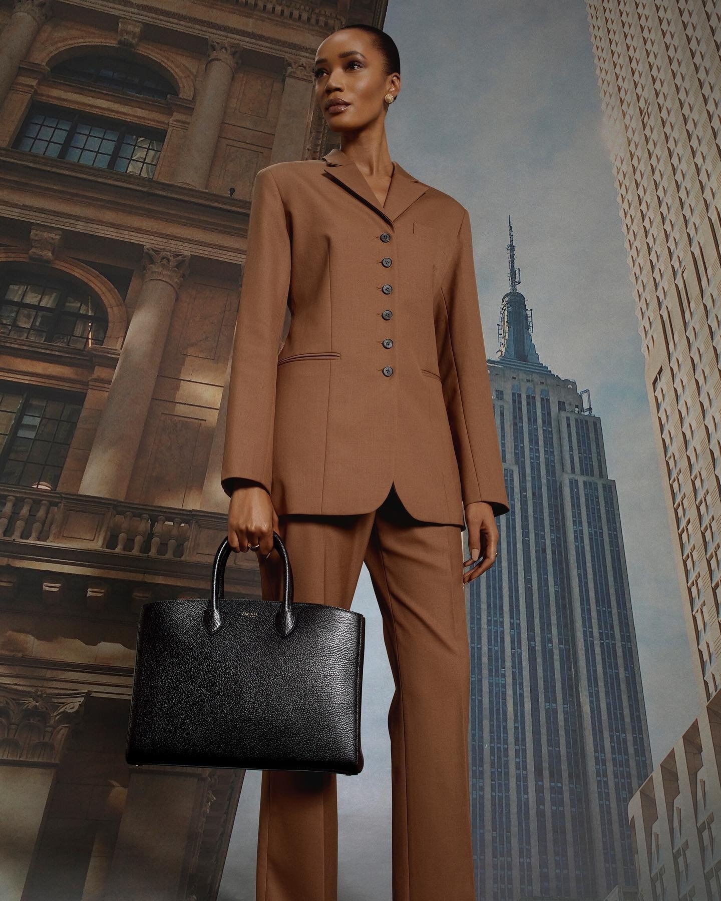 Adult female in a brown business suit holding a black briefcase, posing against an urban backdrop featuring a skyscraper.