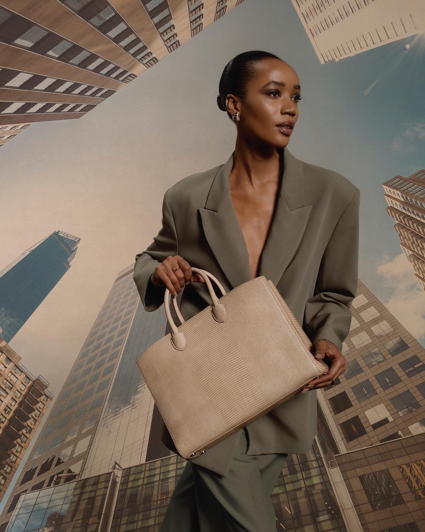Woman in blazer poses in front of a backdrop of tall buildings, holding a light-tan handbag with top handles in a low-angle shot.