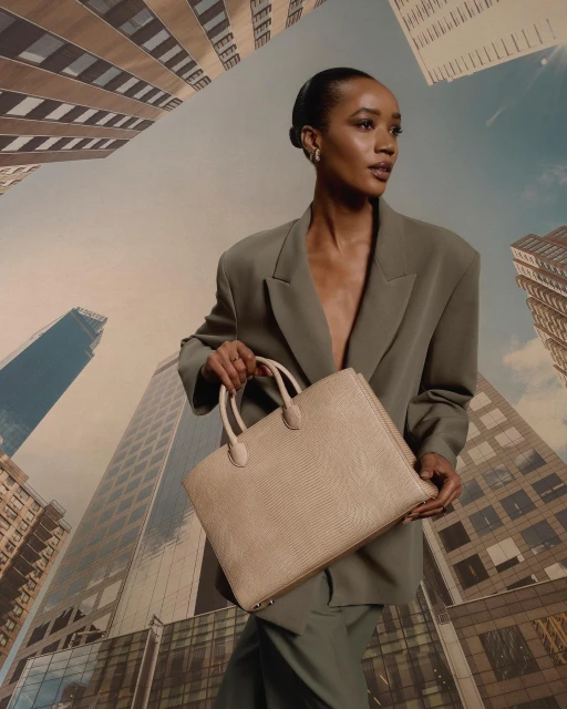 Woman in blazer poses in front of a backdrop of tall buildings, holding a light-tan handbag with top handles in a low-angle shot.