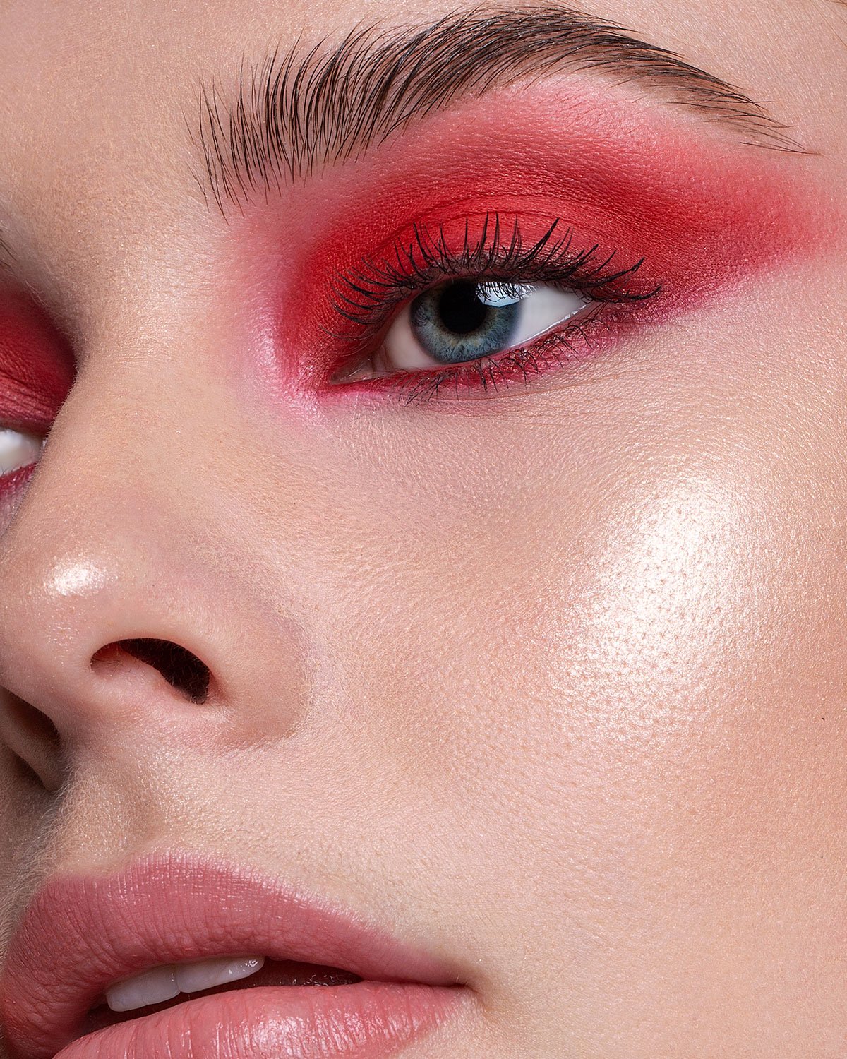 Extreme close-up portrait of a woman featuring bold red eyeshadow, defined brows, and glossy pink lips.