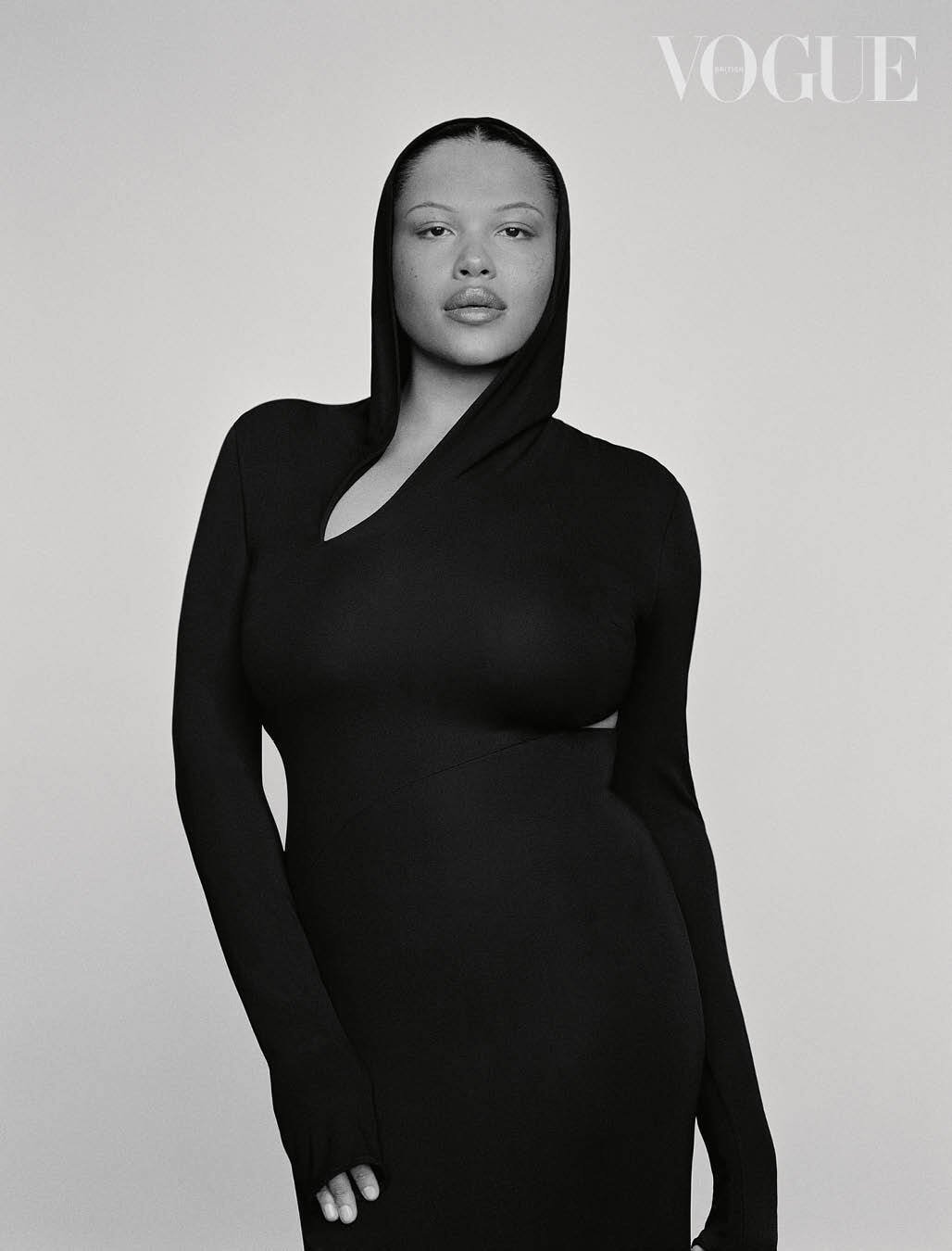 Close-up studio portrait of a woman wearing a black hooded dress with long sleeves, looking directly at the camera. Vogue logo visible.