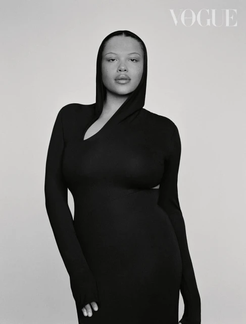 Close-up studio portrait of a woman wearing a black hooded dress with long sleeves, looking directly at the camera. Vogue logo visible.