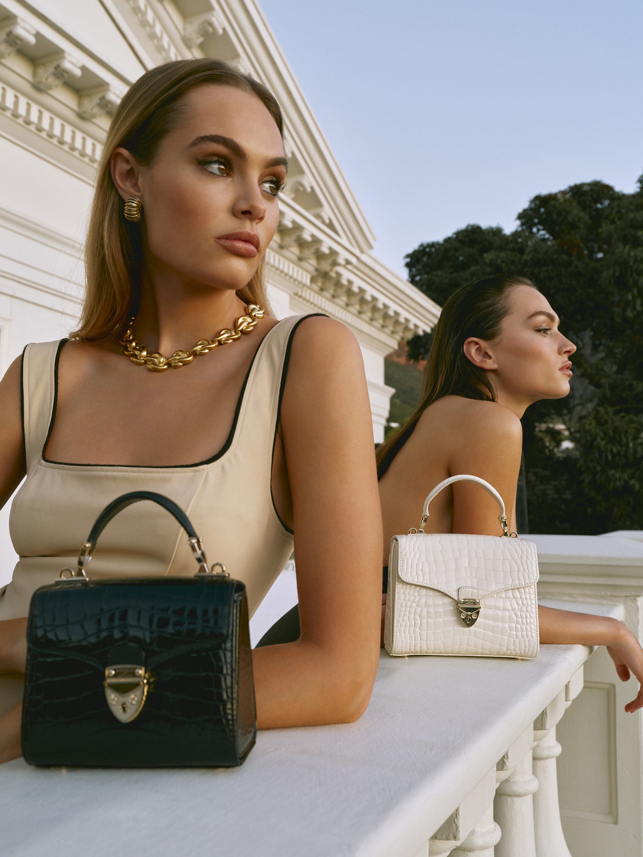 Two women pose with black and white crocodile-skin handbags against a white building and sky backdrop, showcasing luxury fashion.