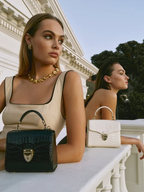 Two women pose with black and white crocodile-skin handbags against a white building and sky backdrop, showcasing luxury fashion.