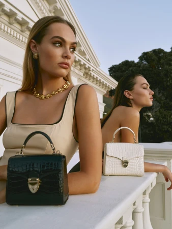 Two women pose with black and white crocodile-skin handbags against a white building and sky backdrop, showcasing luxury fashion.