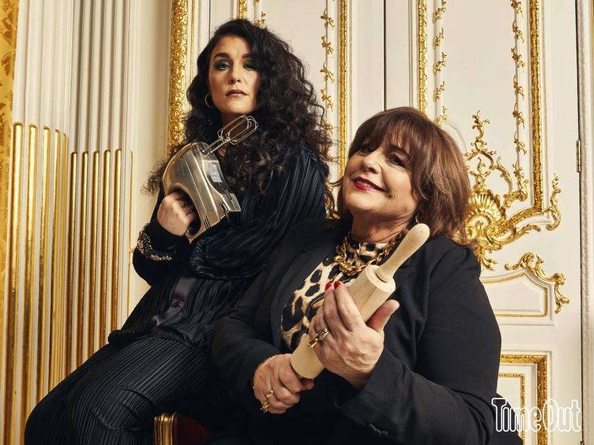 Two women in a photo studio pose with kitchen appliances, including a hand mixer and rolling pin, against a gold decorative wall.