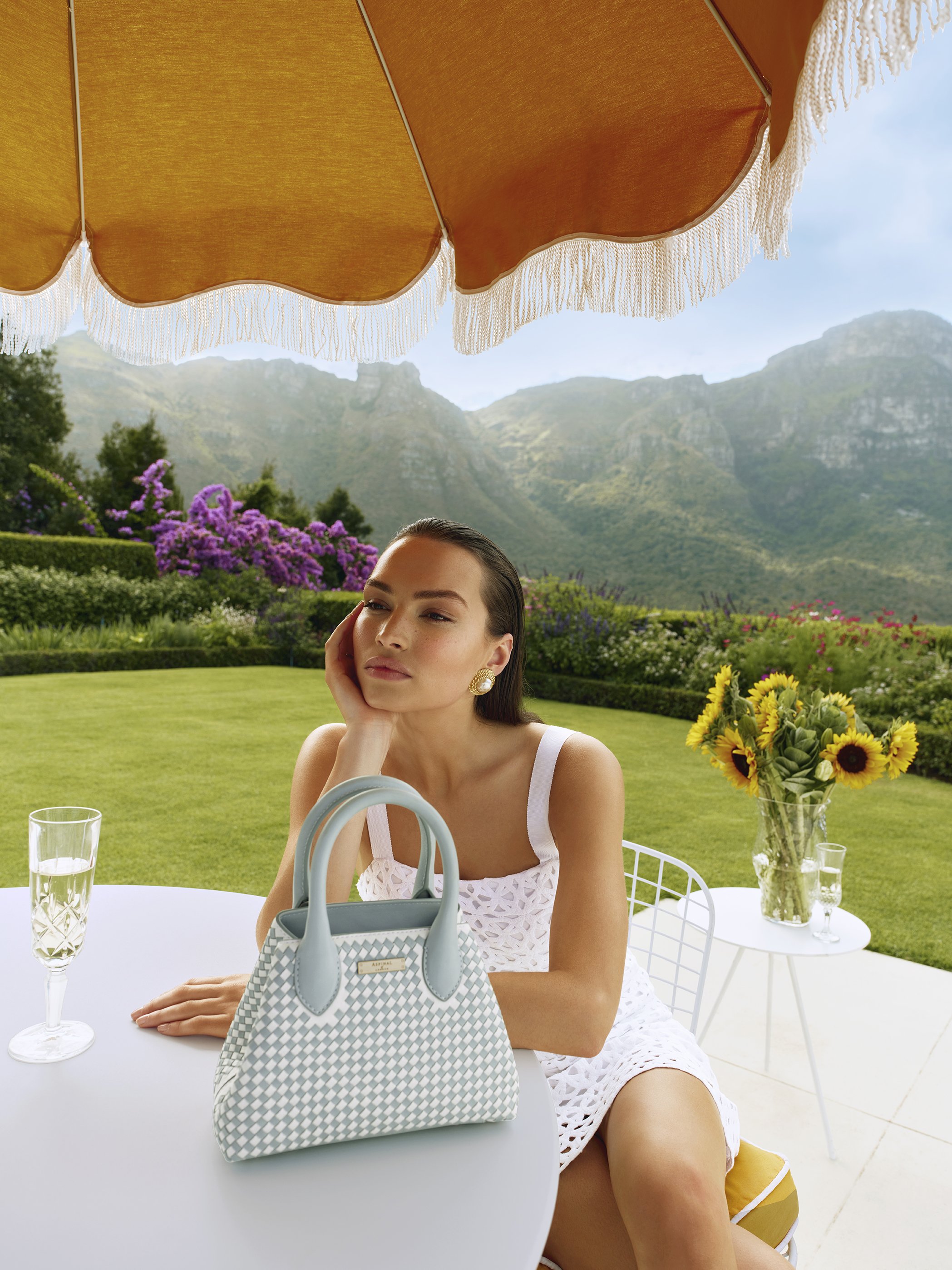 Elegant woman sits at table outdoors with handbag, drink, flowers. Sunny day, mountain backdrop under umbrella.