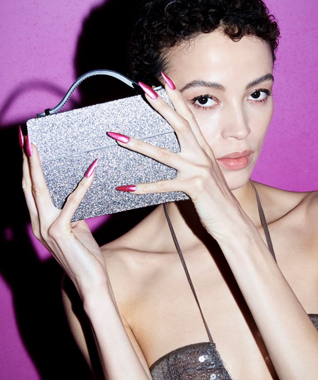 Woman with pink nails holds a silver glitter purse in a studio setting against a vibrant magenta backdrop, fashion and beauty shot.