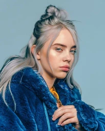 Portrait of Billie Eilish wearing a blue furry jacket with silver-grey hair tied up in a half bun, posed against a pale blue backdrop.