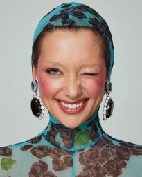 Close-up portrait of a woman winking, wearing a patterned blue hood and large crystal earrings by makeup artist Rachel Singer Clark.