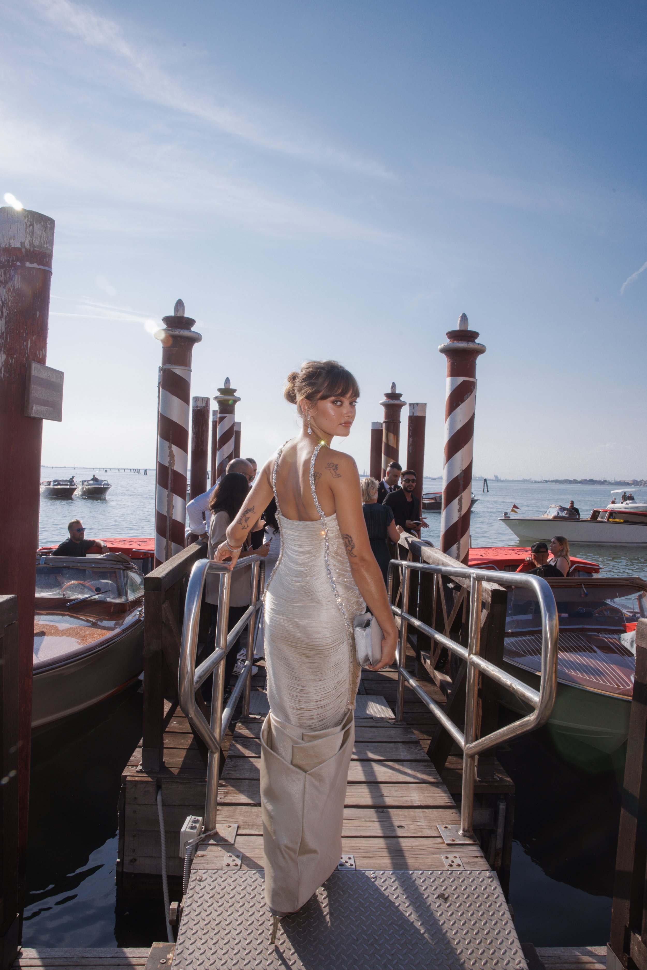 Woman in embellished gown walks a pier in Venice, Italy, with boats and the bright sky creating an elegant fashion moment.