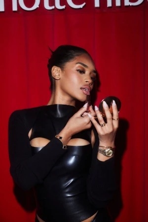 Woman applies lipstick in a close-up shot, wearing a black top against a red background with the Charlotte Tilbury logo visible.