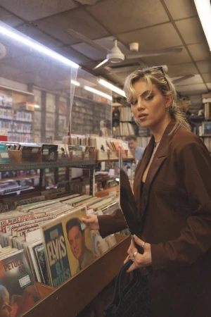 Blonde woman in a brown suit browsing vinyl records at a record store with a clear plastic partition on the record shelf.