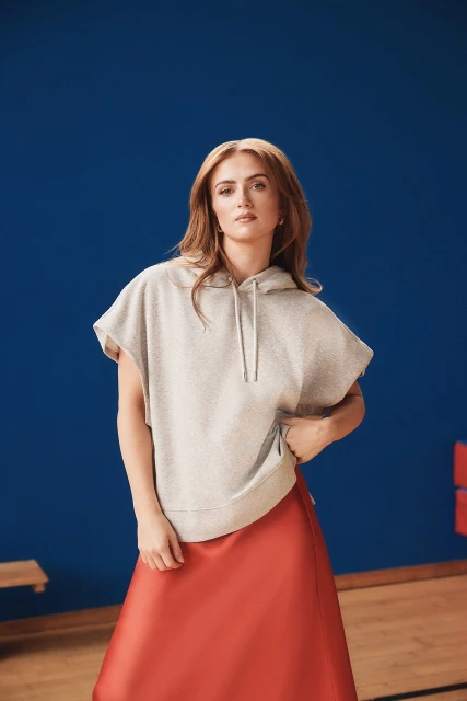 A woman with long red hair poses against a blue background wearing a sleeveless hoodie sweatshirt and a red skirt for fashion.