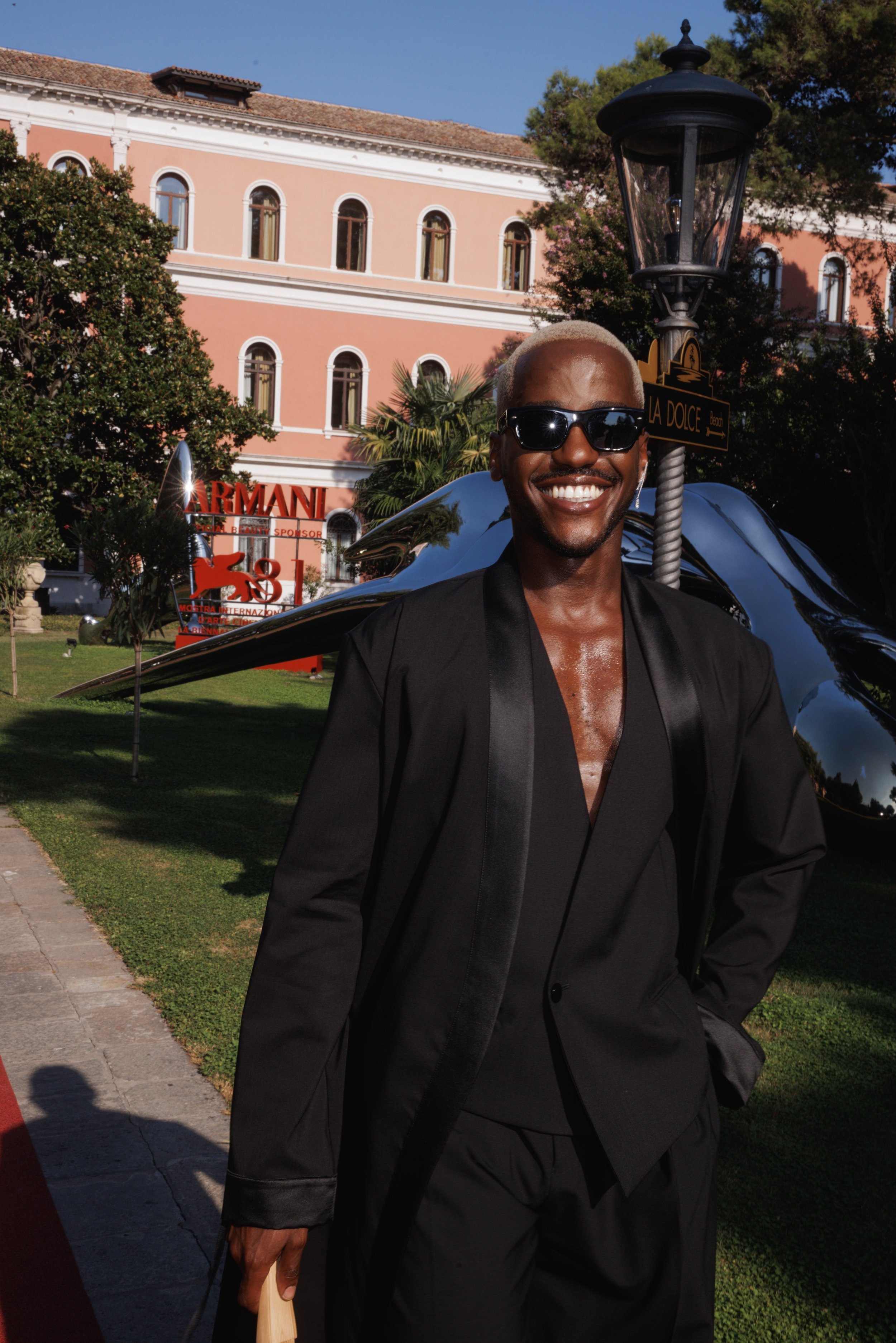 Man in black suit and sunglasses smiles in an outdoor setting, featuring a building, sculpture, and streetlight in the background.