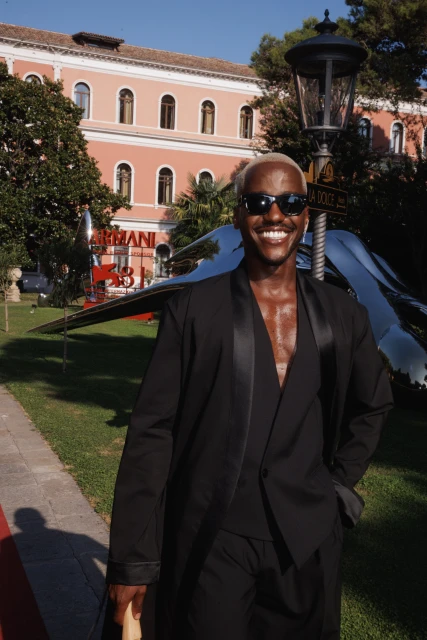 Man in black suit and sunglasses smiles in an outdoor setting, featuring a building, sculpture, and streetlight in the background.
