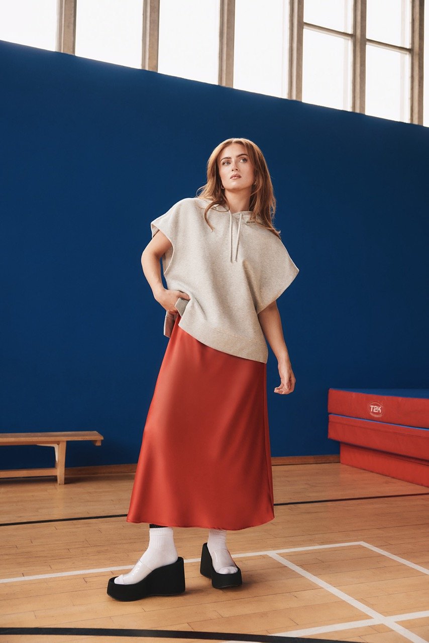 Woman posing in a modern grey hoodie and red skirt, standing on a basketball court, minimalist fashion photography.