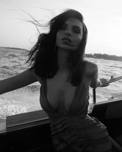 Black and white portrait of an adult woman with wind-blown hair in a low-cut dress on a boat, capturing a moment on the water.