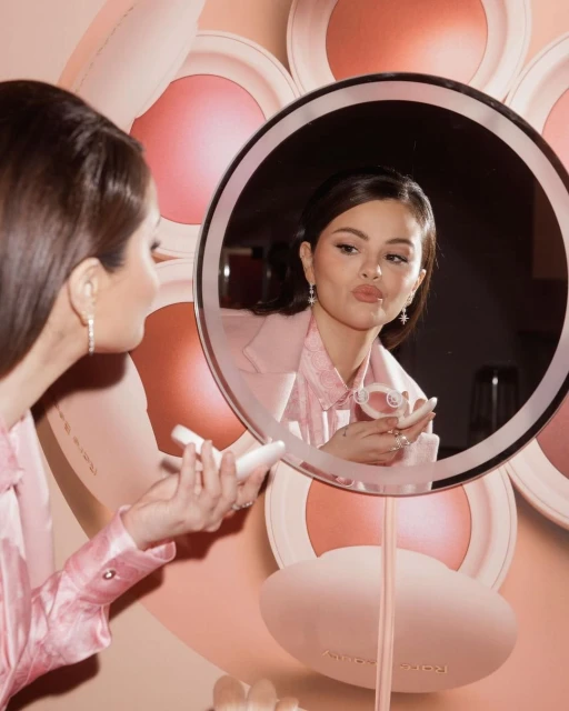 Brunette woman in pink suit pouts and applies makeup in front of round makeup mirror with blush compact display in background.