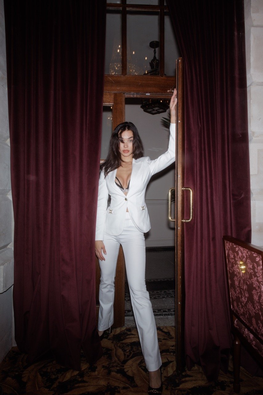 Elegant woman in a white suit poses in a doorway with red velvet curtains. The setting appears to be a high-end restaurant.