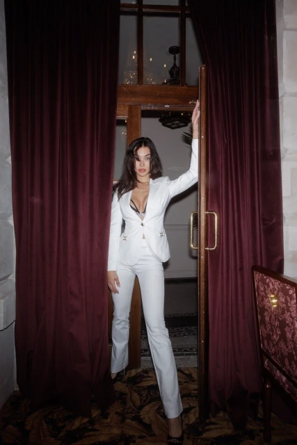 Elegant woman in a white suit poses in a doorway with red velvet curtains. The setting appears to be a high-end restaurant.