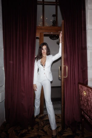 Elegant woman in a white suit poses in a doorway with red velvet curtains. The setting appears to be a high-end restaurant.