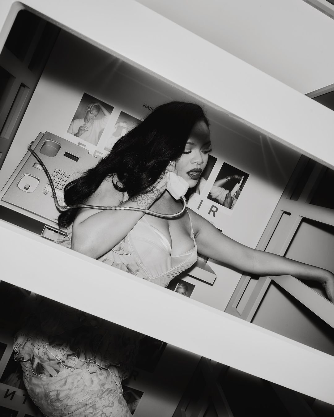 Black and white studio portrait of Rihanna in a telephone booth, holding a vintage telephone receiver to her ear.