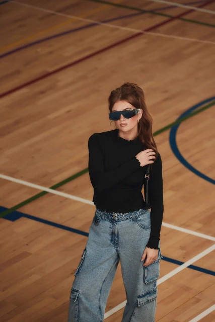 Woman poses in an indoor court wearing a black long-sleeve shirt, baggy cargo jeans, and futuristic sunglasses, showcasing fashion style.