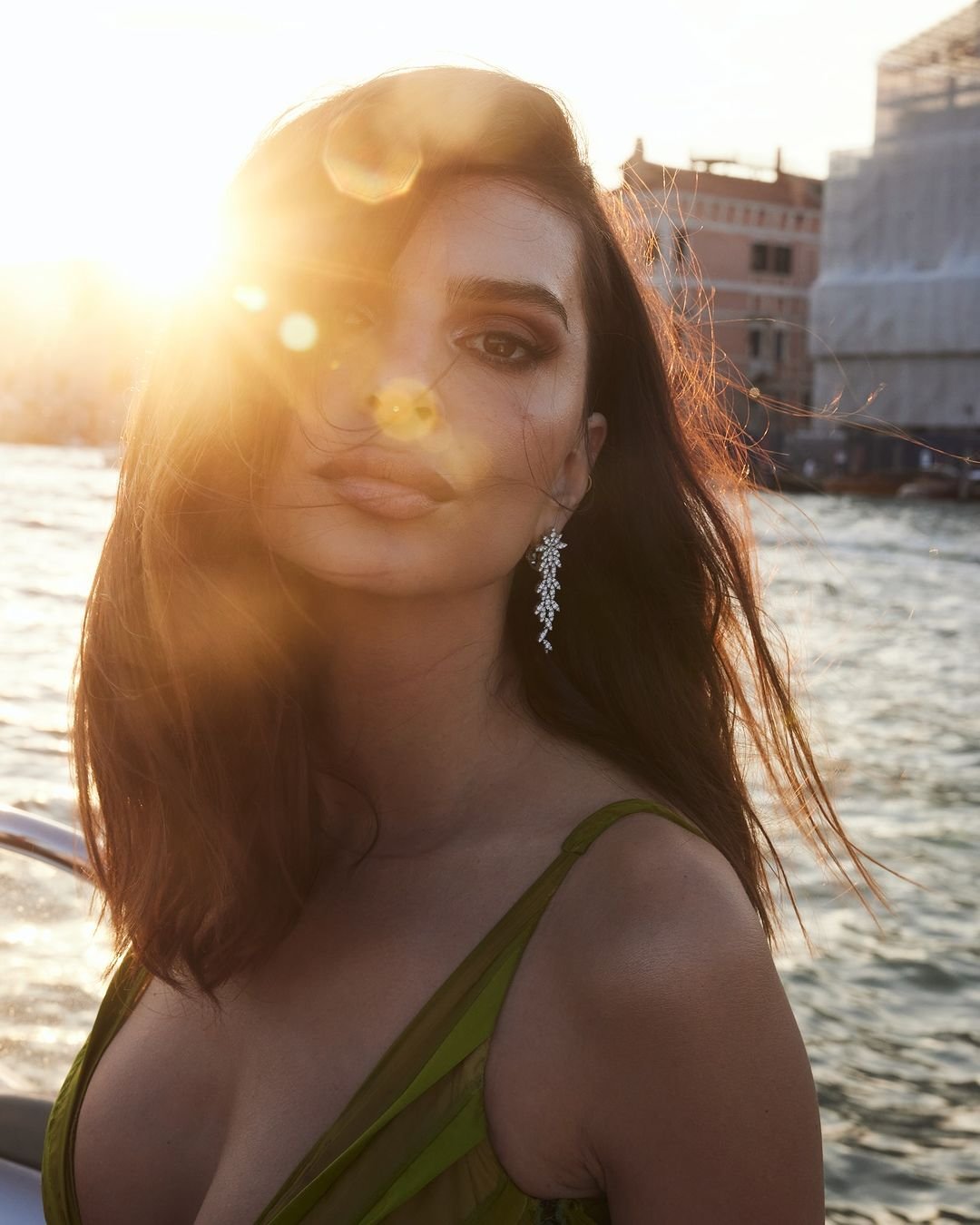 Portrait of a woman with long brown hair wearing earrings and a green dress with Venice buildings and bright sun in the background.