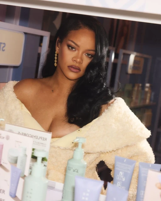 Elegant woman with long hair and neutral makeup poses indoors, wearing a cream-colored furry shawl and gold earrings near beauty products.