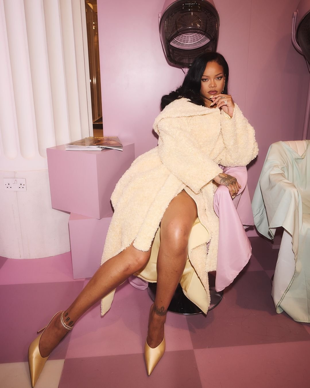 Rihanna poses indoors in a cream robe and gold heels, seated in a pink room with salon equipment. Celebrity fashion and beauty.