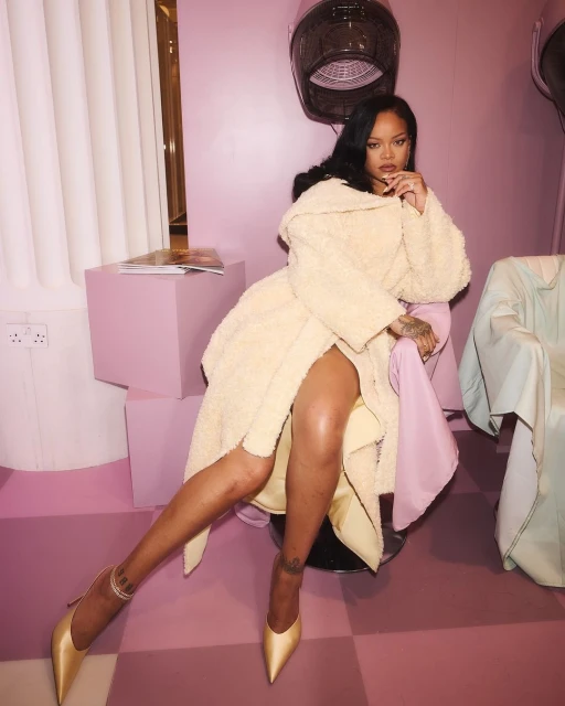 Rihanna poses indoors in a cream robe and gold heels, seated in a pink room with salon equipment. Celebrity fashion and beauty.