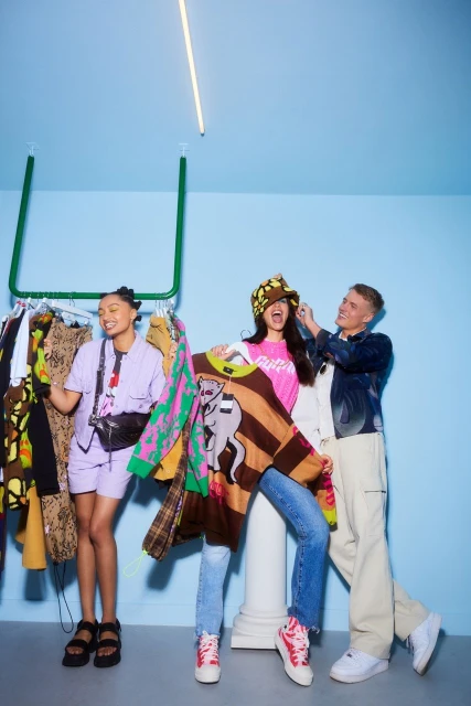 Three people are inside a colorful clothing store holding up outfits. Two adult females and one adult male are the subjects.