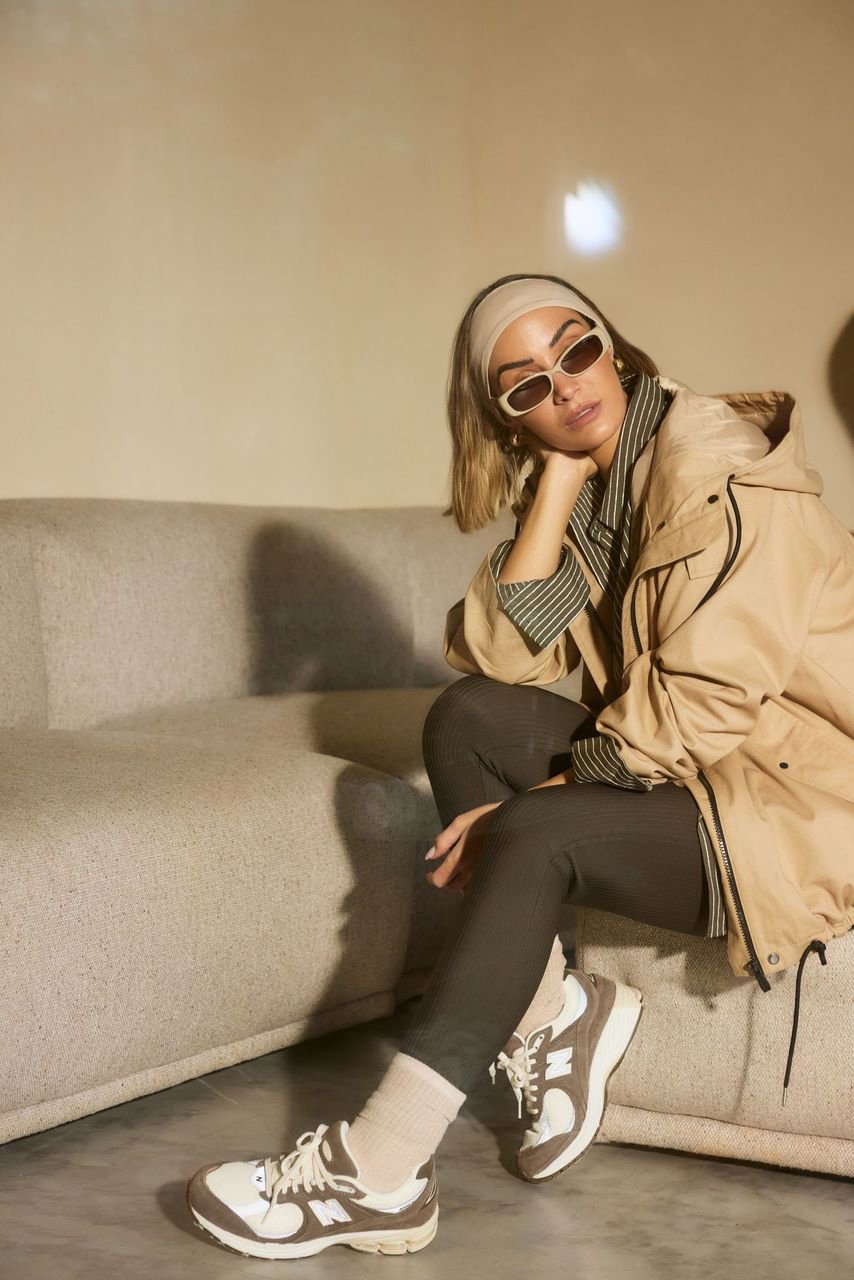 Full shot of an adult woman in a neutral-colored windbreaker, sunglasses, and sneakers sitting on a grey couch indoors.