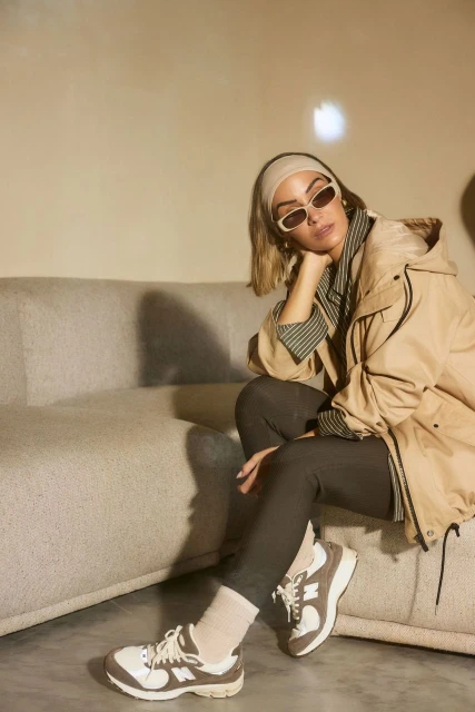 Full shot of an adult woman in a neutral-colored windbreaker, sunglasses, and sneakers sitting on a grey couch indoors.
