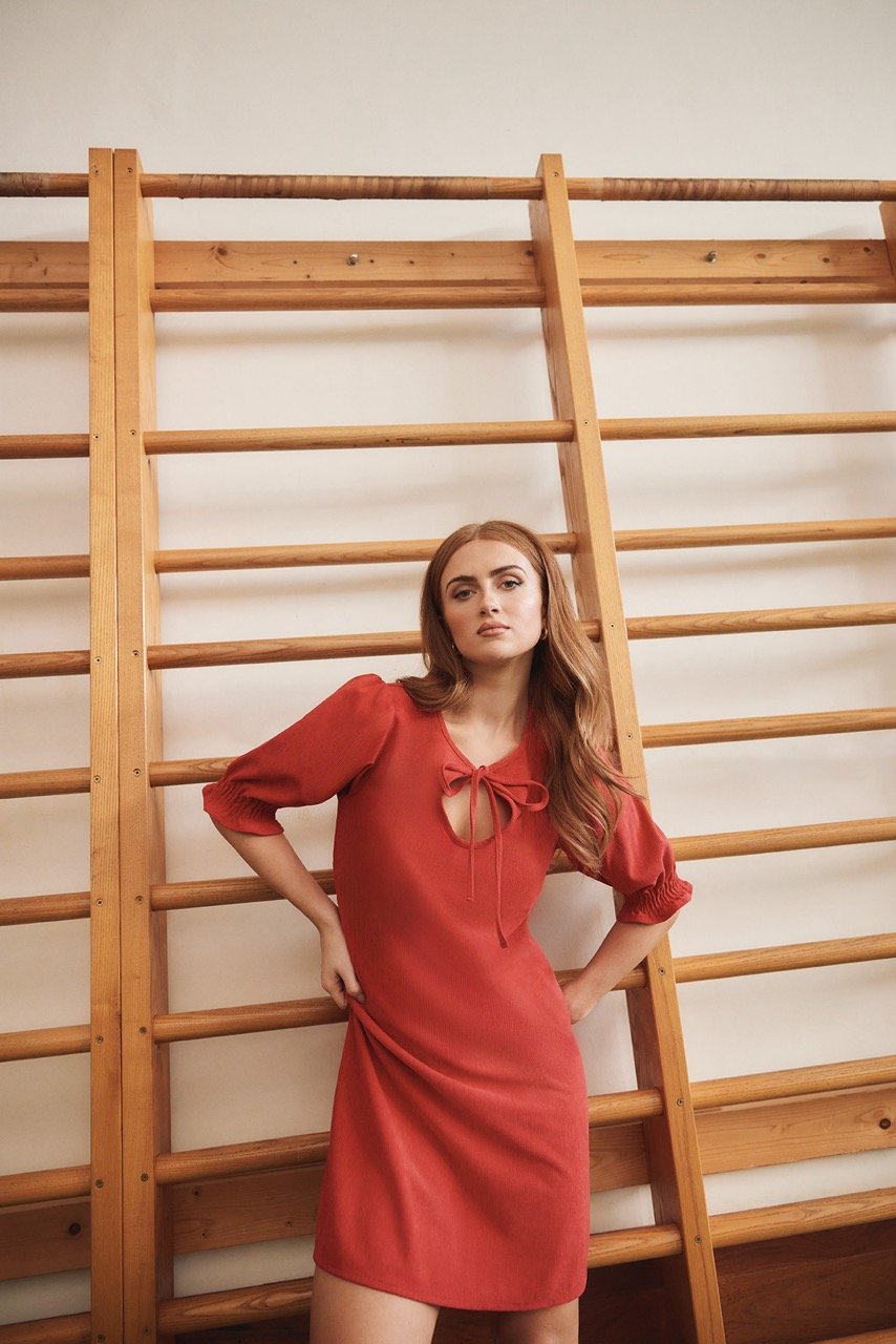 A young fair-skinned woman with long wavy auburn hair, wearing an orange dress, posing in front of a wall-mounted gymnastics ladder.