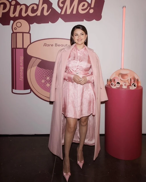 Selena Gomez wearing a light pink dress and coat, smiling in front of a Rare Beauty backdrop and display. Beauty and fashion industry.
