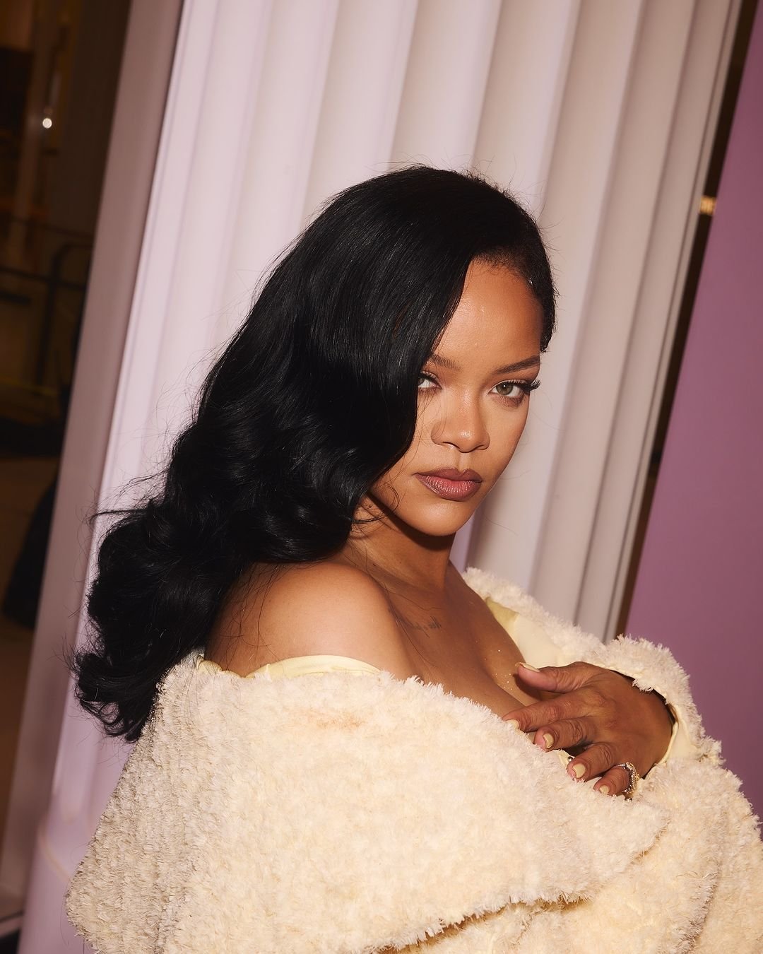 Close-up portrait of Rihanna with long, wavy black hair, wearing a cream-colored, textured coat against a muted pink and white backdrop.