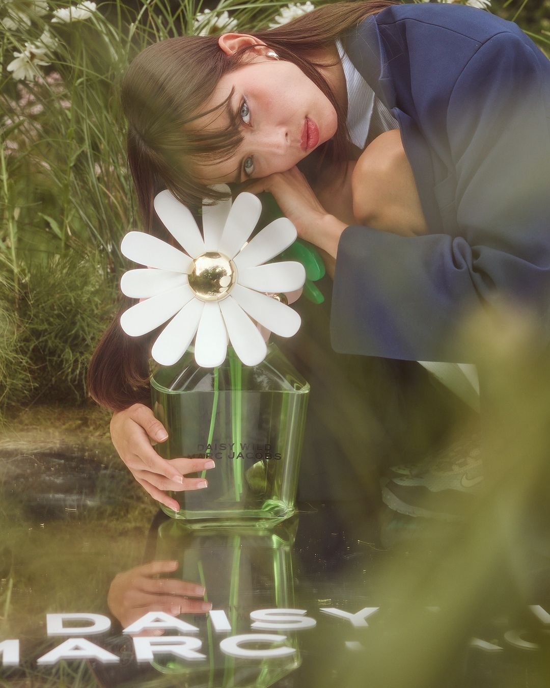 Young woman in blue blazer poses outdoors with large daisy-shaped flower in a glass vase in a field of green grass and white flowers.