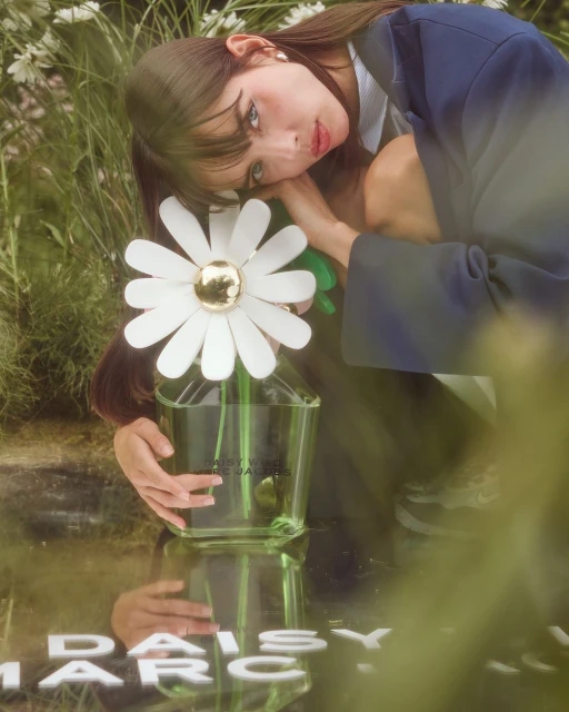 Young woman in blue blazer poses outdoors with large daisy-shaped flower in a glass vase in a field of green grass and white flowers.