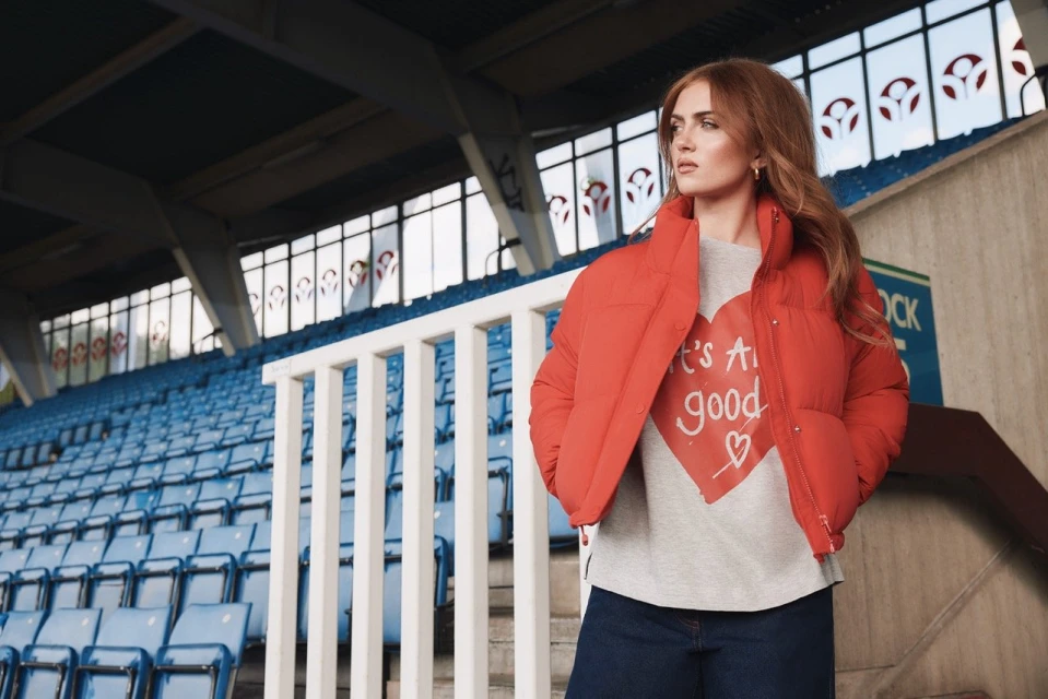 Adult female in an orange puffer jacket poses in stadium seating with blue seats and concrete floors, looking to the upper left.