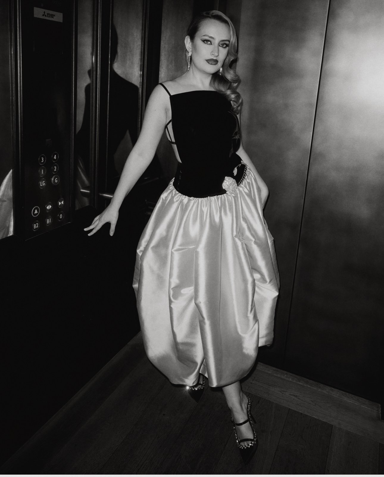 Elegant woman in a black and iridescent dress, posing in an elevator. The photo is black and white, with a dramatic and poised presentation.