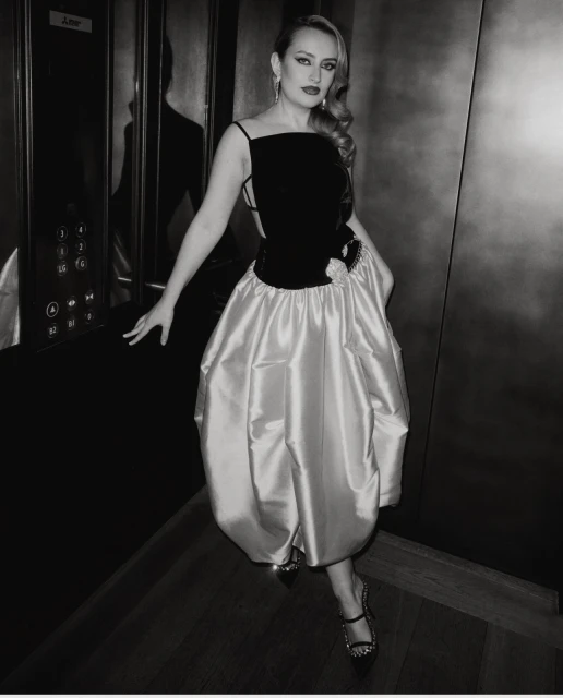 Elegant woman in a black and iridescent dress, posing in an elevator. The photo is black and white, with a dramatic and poised presentation.