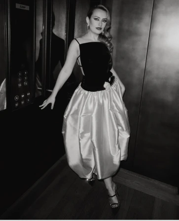 Elegant woman in a black and iridescent dress, posing in an elevator. The photo is black and white, with a dramatic and poised presentation.