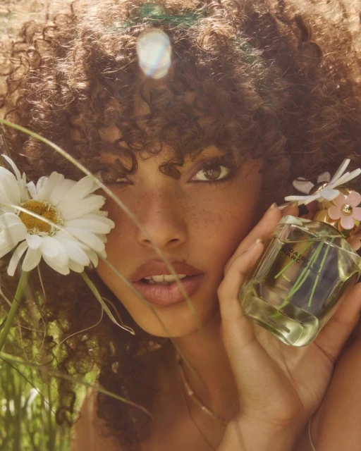 Woman with curly hair holds Daisy Marc Jacobs perfume bottle outdoors among daisies and tall grass in natural light, close-up.