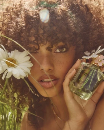 Woman with curly hair holds Daisy Marc Jacobs perfume bottle outdoors among daisies and tall grass in natural light, close-up.